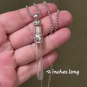 May include: A clear glass vial pendant necklace with a silver-colored metal cap and chain. The vial is 2 inches long. The metal components have decorative patterns. The necklace is designed to hold small items.