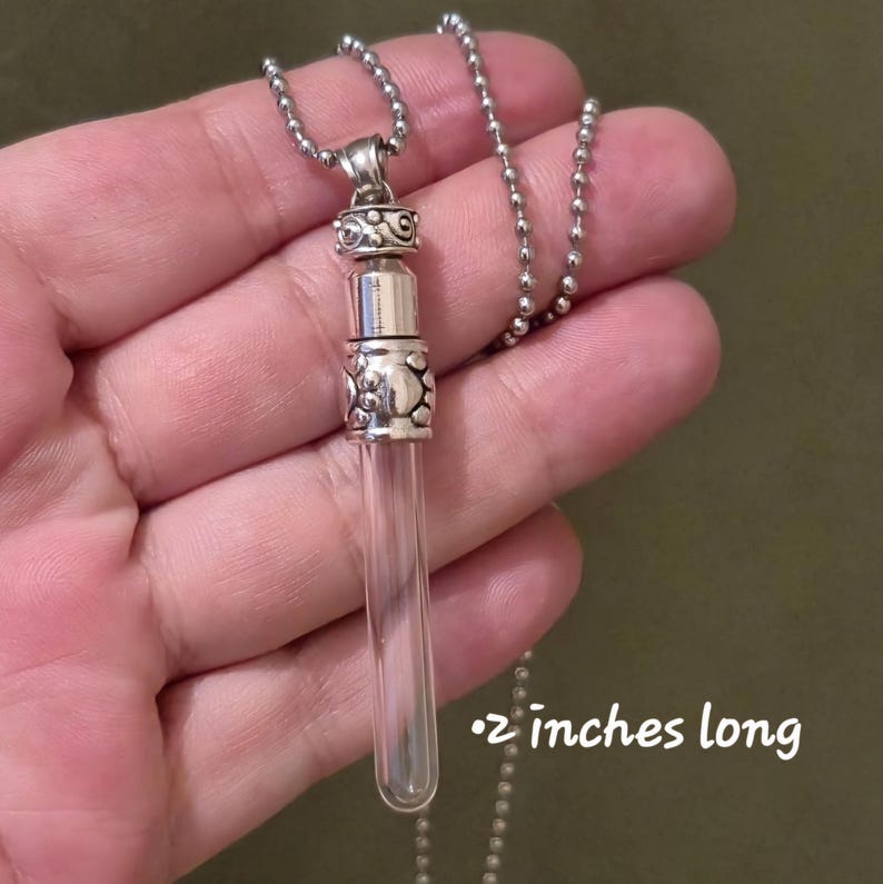 May include: A clear glass vial pendant necklace with a silver-colored metal cap and chain. The vial is 2 inches long. The metal components have decorative patterns. The necklace is designed to hold small items.
