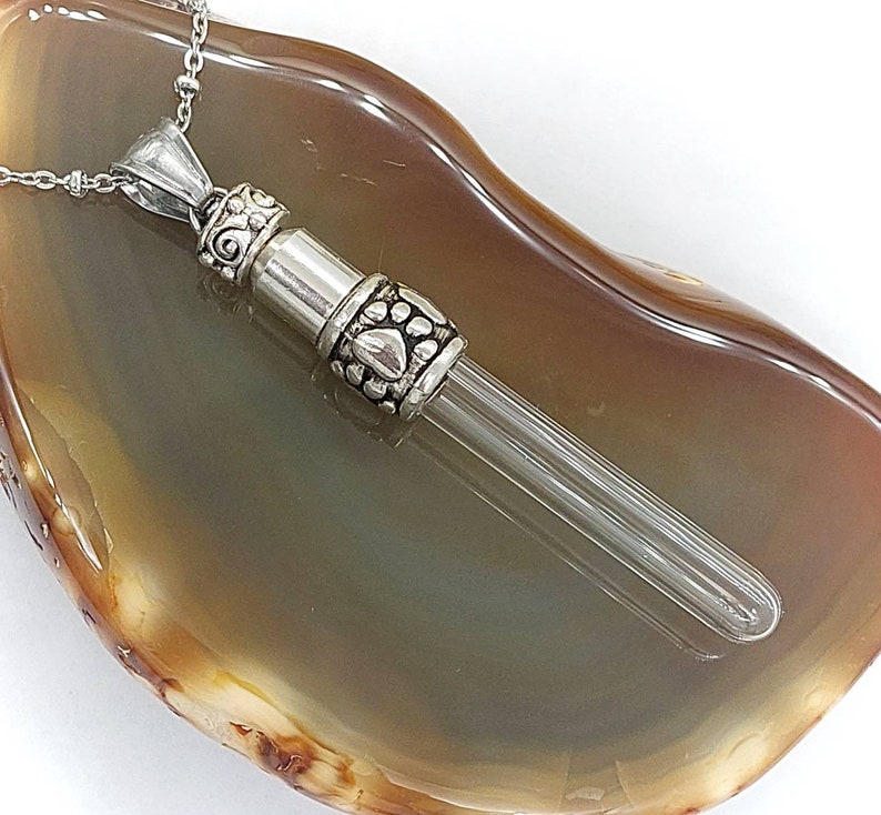 May include: A silver-toned memorial necklace with a clear glass vial pendant. The pendant has decorative paw prints and scrollwork. The necklace rests on a brown and gray agate stone.