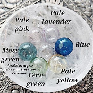 May include: A collection of glass beads in various colors, including pale pink, pale lavender, moss green, fern green, pale yellow, and blue. The beads are arranged in a white bowl. The text "Resolution on your device could cause color variations." is printed below the beads.