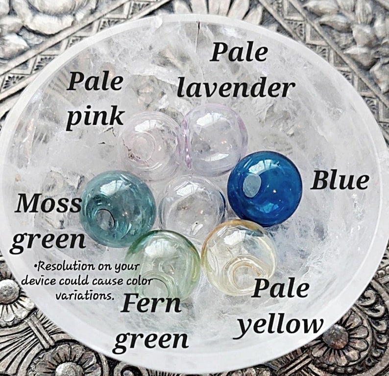 May include: A collection of glass beads in various colors, including pale pink, pale lavender, moss green, fern green, pale yellow, and blue. The beads are arranged in a white bowl. The text "Resolution on your device could cause color variations." is printed below the beads.