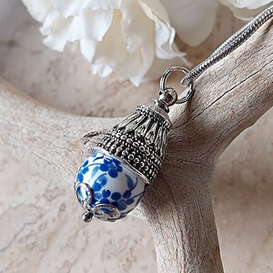 Puede incluir: Un collar con un colgante de plata y un dije de porcelana azul y blanco. El dije presenta un dise&ntilde;o floral y est&aacute; acentuado con detalles plateados.