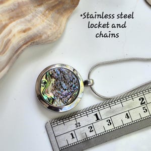 May include: A round stainless steel locket pendant with an iridescent abalone shell inlay, attached to a silver-toned snake chain. A metal ruler shows the locket diameter is approximately 1 inch. Text reads: Stainless steel locket and chains.