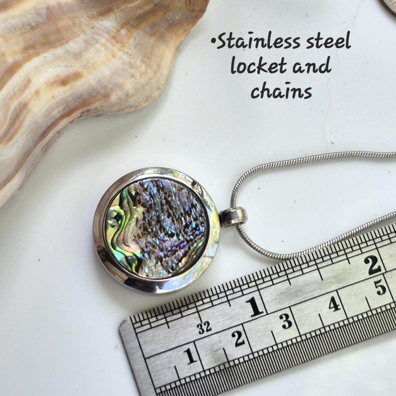 May include: A round stainless steel locket pendant with an iridescent abalone shell inlay, attached to a silver-toned snake chain. A metal ruler shows the locket diameter is approximately 1 inch. Text reads: Stainless steel locket and chains.