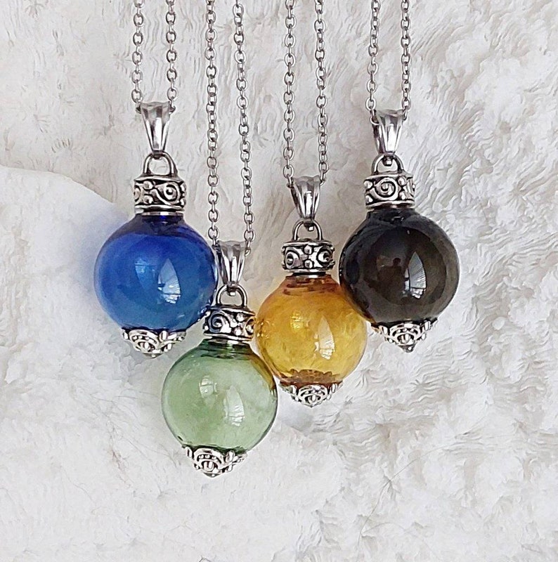 May include: Four glass orb pendants in blue, green, yellow, and dark brown, suspended from silver chains. Each orb has a decorative silver cap and base. The pendants are displayed against a white, textured background.