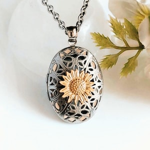 May include: Silver-tone oval locket necklace with a gold-colored sunflower design. The locket features intricate cut-out patterns and hangs from a matching chain. A floral accent is visible in the background.