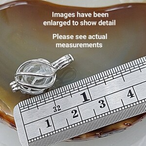 May include: A silver charm in the shape of a cage with a clear crystal inside. The charm is measured against a ruler, showing it is approximately 1 inch in diameter.