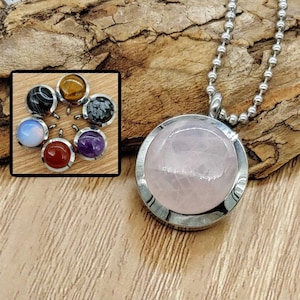 Peut inclure: Un collier en chaîne argentée avec un pendentif rond orné d'une pierre de quartz rose. Le pendentif est doté d'une bélière en argent et d'une finition lisse et polie. Le collier est présenté sur une surface en bois.