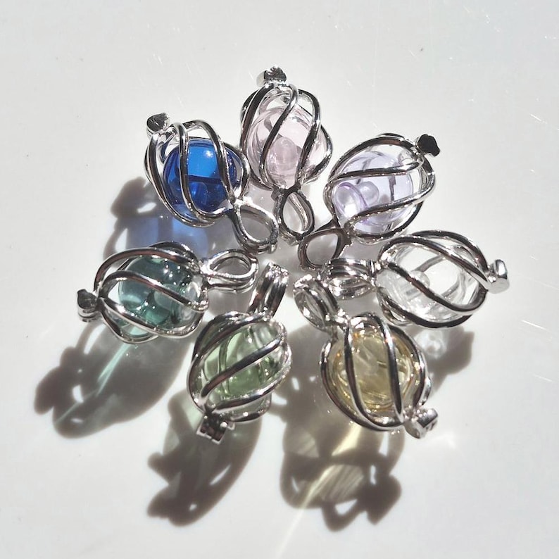 Puede incluir: Una colecci&oacute;n de siete colgantes plateados, cada uno con una gema esf&eacute;rica de colores encerrada en una jaula de alambre. Las gemas son de varios colores, incluyendo azul, rosa, verde y amarillo. Los colgantes est&aacute;n dispuestos en un patr&oacute;n circular sobre una superficie blanca.