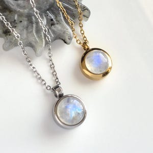 May include: Two necklaces featuring round pendants with iridescent moonstone cabochons. One pendant is set in a polished silver-tone metal casing on a matching chain, while the other is set in a gold-tone metal casing on a matching gold-tone chain.