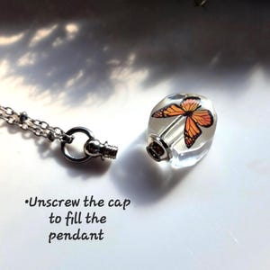 May include: A clear glass pendant containing an orange and black monarch butterfly graphic, attached to a silver-toned metal chain. The pendant features a removable screw-top cap. Text on the image reads: Unscrew the cap to fill the pendant.
