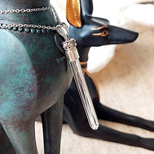 May include: A silver chain necklace with a clear glass pendant containing a bundle of silver wires. The pendant has a silver cap with an ornate design. The necklace is hanging from a statue of an Egyptian god.