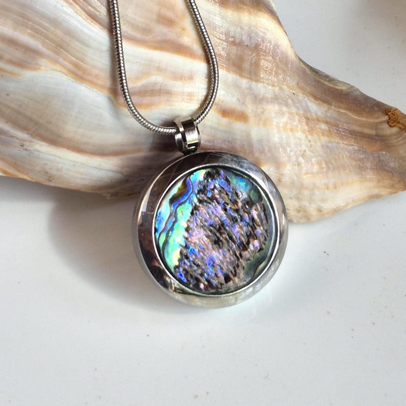 May include: A circular pendant necklace featuring a polished silver-tone metal frame surrounding a vibrant, iridescent abalone shell inlay with swirling patterns of blue, green, and purple, suspended from a matching silver-tone snake chain.