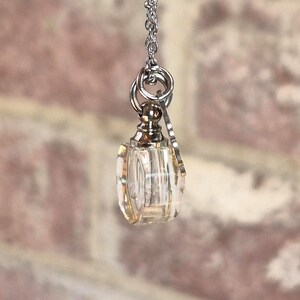 May include: A small, clear glass vial pendant suspended from a silver-toned metal chain. The vial features a metallic silver cap and a small, flat, circular silver charm attached to the jump ring, creating a delicate necklace design.