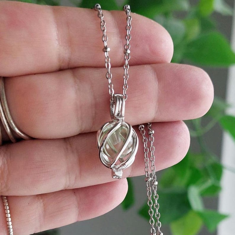 Puede incluir: Un collar plateado con un colgante con dise&ntilde;o de jaula. Dentro de la jaula hay una piedra ovalada de color verde claro. El collar est&aacute; sostenido por una mano, con un fondo verde borroso.