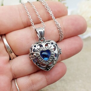 May include: A silver heart-shaped locket necklace with a blue crystal in the center. The locket has an intricate design and is open to reveal a small space inside.