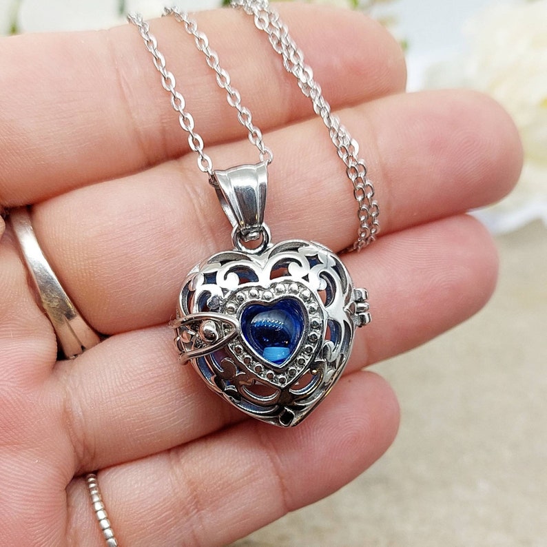 May include: A silver heart-shaped locket necklace with a blue crystal in the center. The locket has an intricate design and is open to reveal a small space inside.
