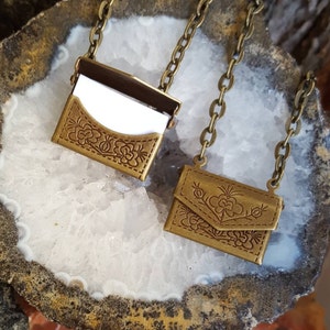 May include: Two gold-toned metal necklaces with small, ornate, floral-embossed, hinged, envelope-shaped pouches. The pouches are open to reveal a small, white, rectangular insert.