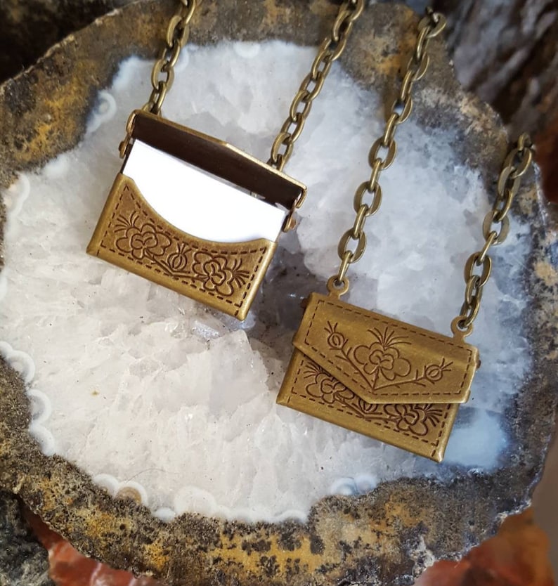 May include: Two gold-toned metal necklaces with small, ornate, floral-embossed, hinged, envelope-shaped pouches. The pouches are open to reveal a small, white, rectangular insert.