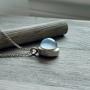 May include: A silver chain necklace with a pendant featuring a round, light blue glass orb encased in a silver frame.