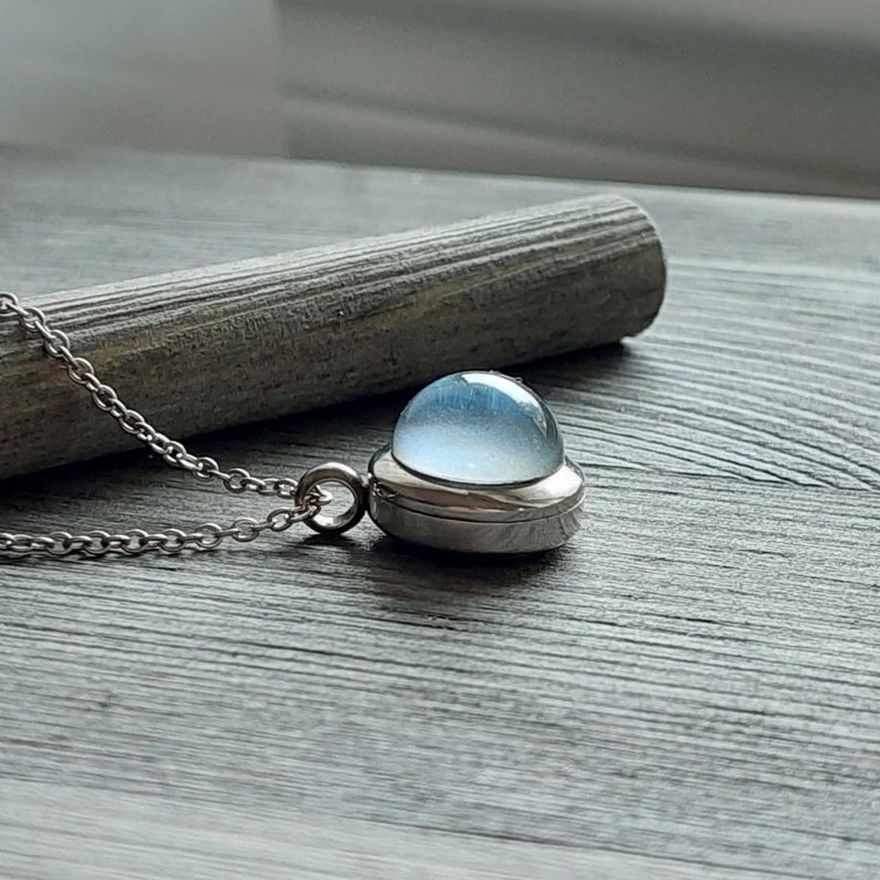 May include: A silver chain necklace with a pendant featuring a round, light blue glass orb encased in a silver frame.