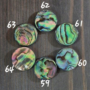 May include: Six circular abalone shell buttons featuring iridescent swirls of green, pink, and purple. Each button is labeled with a white number: 59, 60, 61, 62, 63, and 64. The buttons display natural, unique patterns with a smooth, polished finish.