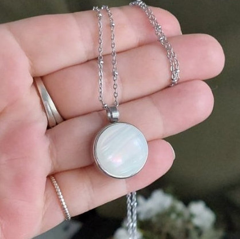 Peut inclure: Un collier argent&eacute; avec un pendentif rond et iris&eacute;. Le pendentif pr&eacute;sente un motif tourbillonnant blanc nacr&eacute; et bleu clair. La cha&icirc;ne du collier est d&eacute;licate et comporte de petites perles rondes. Le bijou est pr&eacute;sent&eacute; sur une main.