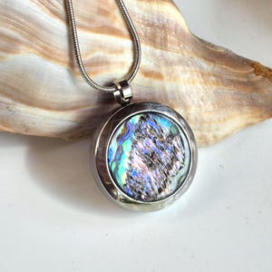 May include: A round silver-toned pendant necklace featuring a circular inlay of iridescent abalone shell with swirling blue, green, and purple hues. The pendant is suspended from a matching silver-toned snake chain, showcasing a polished metal frame.
