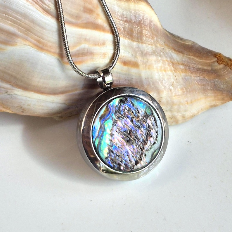 May include: A round silver-toned pendant necklace featuring a circular inlay of iridescent abalone shell with swirling blue, green, and purple hues. The pendant is suspended from a matching silver-toned snake chain, showcasing a polished metal frame.