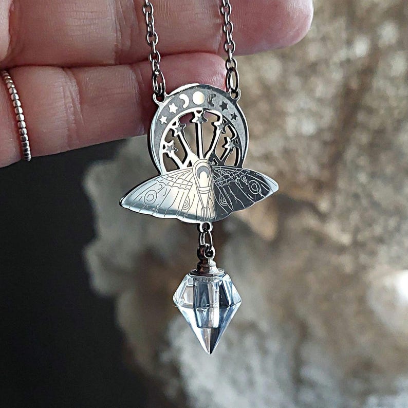 May include: A silver-toned metal pendant necklace featuring a moth design with intricate wing patterns, topped by a crescent moon and star cutout motif. A clear, faceted, diamond-shaped crystal drop hangs from the base of the moth pendant.