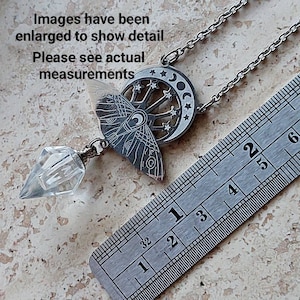 May include: A silver-toned necklace featuring a moth pendant with a crescent moon and star cutout design, attached to a clear, faceted crystal point. A metal ruler is visible, showing the pendant length is approximately 2.5 inches long.