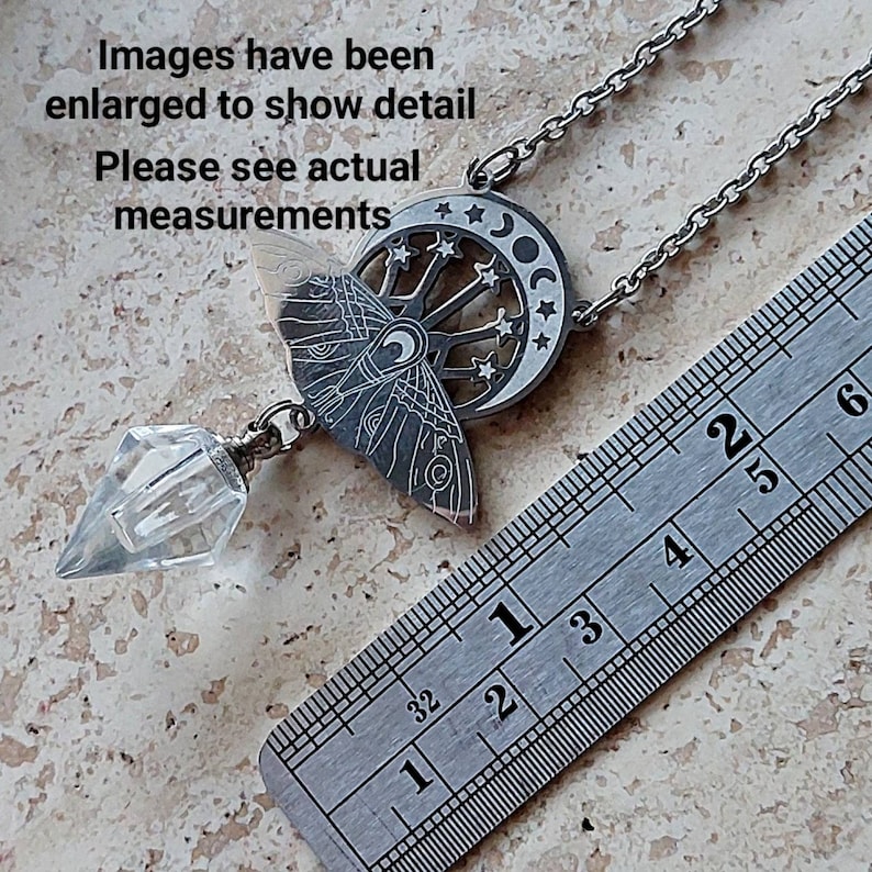 May include: A silver-toned necklace featuring a moth pendant with a crescent moon and star cutout design, attached to a clear, faceted crystal point. A metal ruler is visible, showing the pendant length is approximately 2.5 inches long.