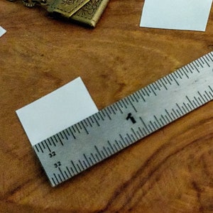 May include: A white square piece of paper is measured with a silver ruler. The ruler is marked in inches and has the number "1" visible.