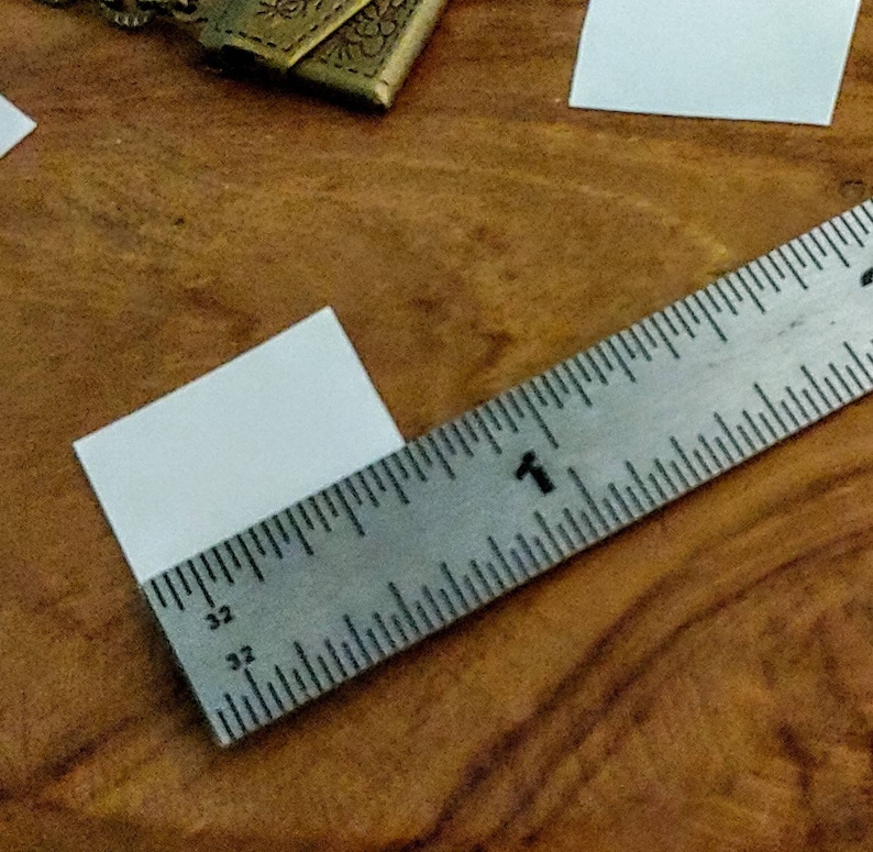 May include: A white square piece of paper is measured with a silver ruler. The ruler is marked in inches and has the number "1" visible.