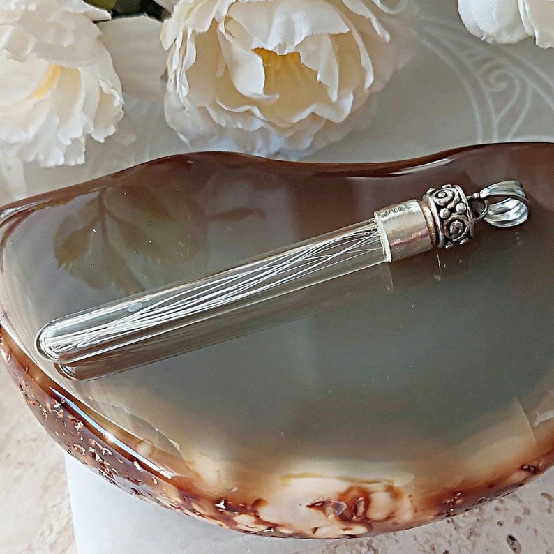 May include: A clear glass tube pendant with a silver cap and intricate detailing. The tube is filled with thin, white fibers. The pendant is resting on a brown agate slice.