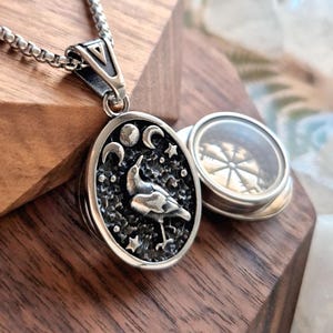 May include: Silver-toned pendant necklace featuring an oval locket with a bird design, moons, and stars. The locket opens to reveal a compass design. The necklace is displayed on a wooden surface.