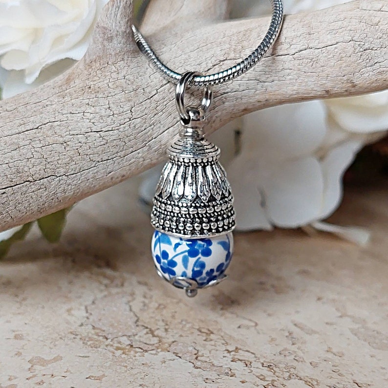 Puede incluir: Un collar de plata con un colgante de porcelana azul y blanco con un dise&ntilde;o floral. El colgante est&aacute; coronado con una tapa de plata con detalles intrincados.