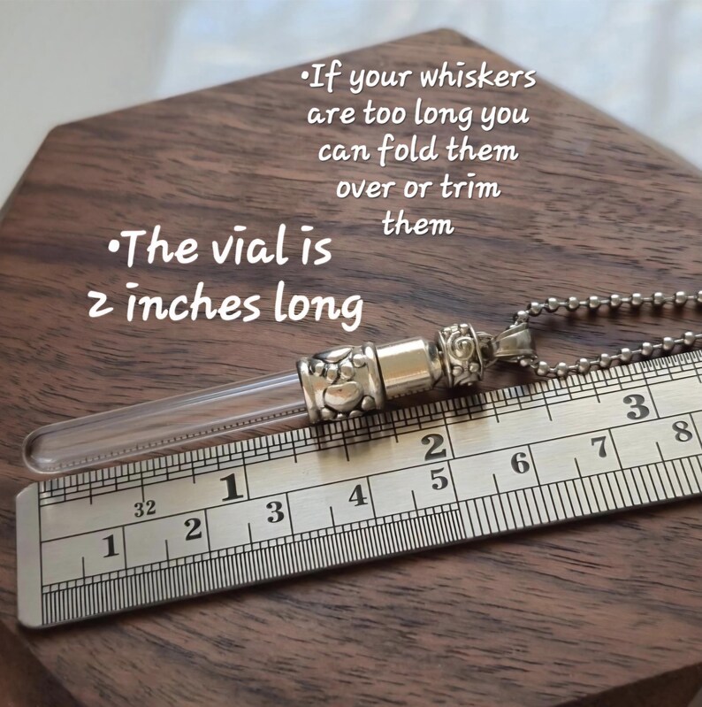 May include: A clear glass vial pendant with silver-colored metal accents, resting on a wooden surface next to a ruler. The vial is 2 inches long. Text on the image reads: "If your whiskers are too long you can fold them over or trim them."