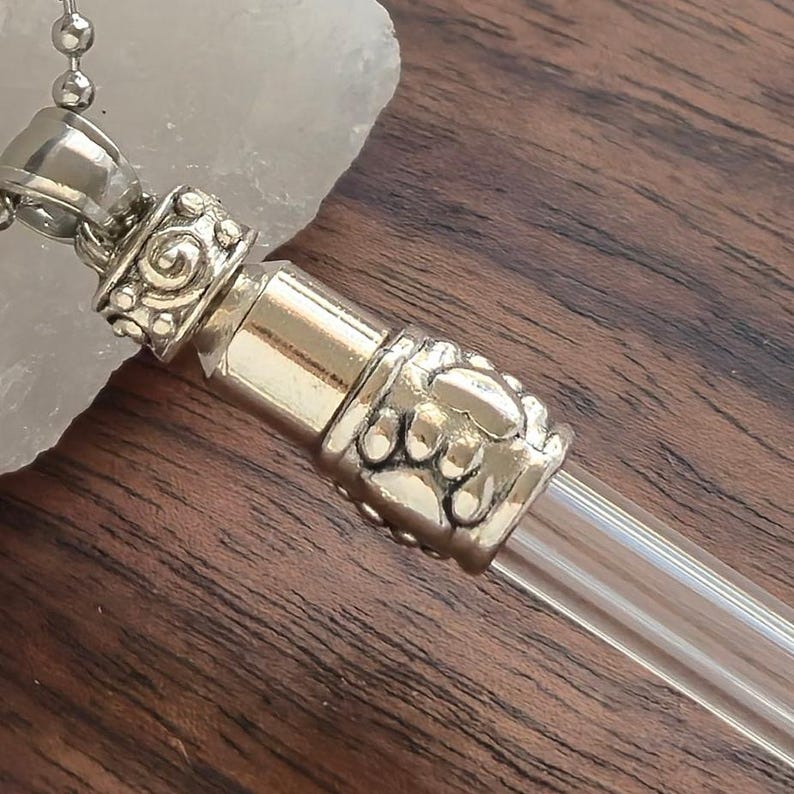 May include: A silver-toned pendant with a clear glass vial. The pendant has ornate detailing and a decorative cap. The vial is cylindrical and appears to be empty. The pendant hangs from a silver chain.
