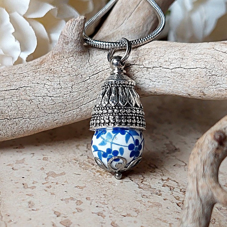 Puede incluir: Un collar de plata con un colgante en forma de bola de estilo porcelana azul y blanco. El colgante tiene un dise&ntilde;o de filigrana de plata alrededor de la bola y una tapa de plata en la parte superior.