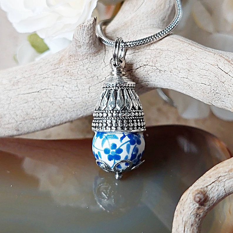 Puede incluir: Un collar con un colgante de plata y una cuenta de porcelana azul y blanca. La cuenta tiene un dise&ntilde;o floral y est&aacute; coronada con una tapa de plata.