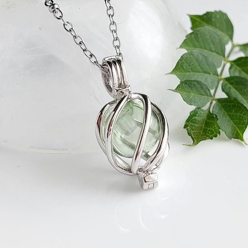 Puede incluir: Un collar con colgante plateado con un dise&ntilde;o de jaula que encierra una piedra esf&eacute;rica verde claro. El collar tiene una cadena fina. El fondo es blanco con hojas verdes.