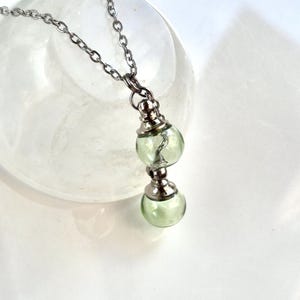 May include: A silver-toned necklace featuring a pendant with two clear, round, green glass orbs. The orbs are connected by a silver-toned metal piece. The necklace chain is also silver-toned. The pendant has a small, silver-toned figure inside the top orb.