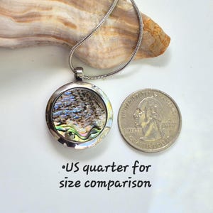 May include: A round silver-toned pendant necklace featuring an iridescent abalone shell inlay. The pendant is displayed next to a US quarter for size comparison, with the text US quarter for size comparison printed below the coin.