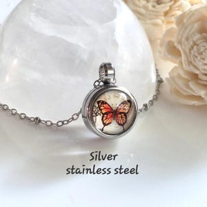 Puede incluir: Collar con colgante de acero inoxidable plateado. El medall&oacute;n redondo presenta un dise&ntilde;o de mariposa con alas naranjas y rojas. El collar tiene una cadena plateada.