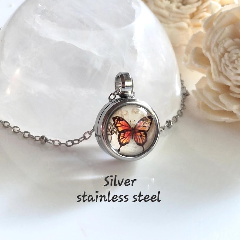 Puede incluir: Collar con colgante de acero inoxidable plateado. El medall&oacute;n redondo presenta un dise&ntilde;o de mariposa con alas naranjas y rojas. El collar tiene una cadena plateada.