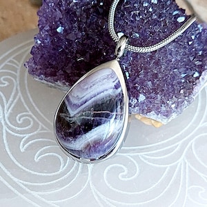 May include: A silver chain necklace with a teardrop-shaped pendant made of amethyst. The amethyst is a deep purple color with white streaks throughout.