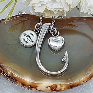 May include: A silver-colored necklace featuring a large fish hook pendant, a heart-shaped charm, and a round charm with a fish illustration and the words "GOAL FISHING". The necklace is displayed on a decorative surface.
