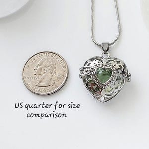 May include: Silver heart-shaped locket necklace with a green glass charm inside. The locket is open and the charm is visible. A US quarter is shown for size comparison.