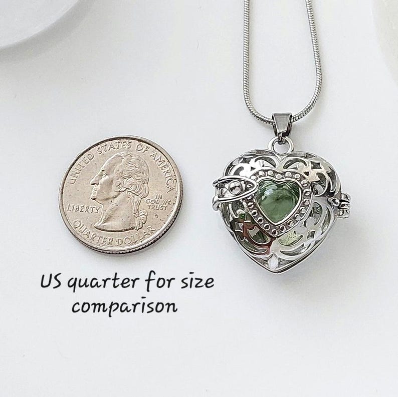 May include: Silver heart-shaped locket necklace with a green glass charm inside. The locket is open and the charm is visible. A US quarter is shown for size comparison.